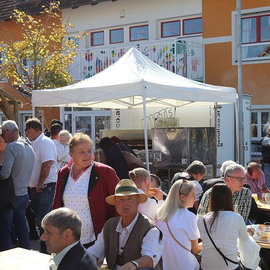 Eine Gruppe von Menschen versammelt sich vor einem Gebäude. Eine Frau steht unter einem weißen Zelt. Ein Mann mit Hut sitzt mit anderen an einem Tisch.