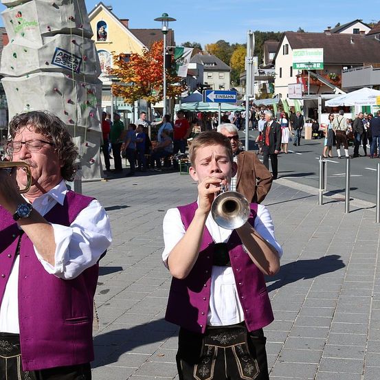 Zwei Männer in traditioneller deutscher Kleidung spielen Trompeten auf einer belebten Straße, mit Gebäuden, einer Kletterwand und einem geschäftigen Markt im Hintergrund.