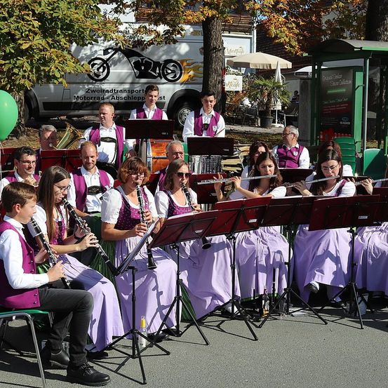 Eine Gruppe von Musikern, gekleidet in lila Westen, spielt auf der Straße mit verschiedenen Instrumenten, darunter Flöten und Trompeten, während ein grüner Ballon in der Nähe schwebt.