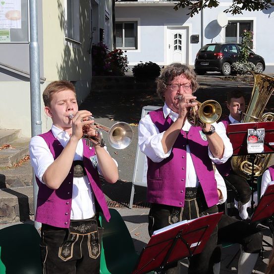 Eine Gruppe von Musikern in traditioneller deutscher Kleidung spielt im Freien. Ein junger Junge spielt Trompete, während ein Mann hinter ihm Posaune spielt. Ein anderer Mann spielt Tuba. Sie stehen auf einem Betonpfad.