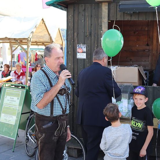 Ein älterer Mann in traditioneller bayerischer Kleidung spricht in ein Mikrofon. In der Nähe beobachten zwei Jungen und ein erwachsener Mann. Hinter ihnen sind grüne Luftballons an einem Holzhäuschen befestigt.