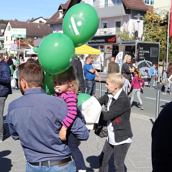 Mehrere Menschen gehen auf einer belebten Straße, ein Mann trägt ein Kind mit grünen Luftballons. Eine Frau reicht dem Kind eine Papiertüte. Im Hintergrund stehen mehrere Gebäude und Zelte.