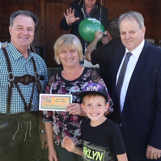 Eine Familie mit einem Kind, die gemeinsam mit einem anderen Mann steht und alle lächelt. Ein Mann trägt traditionelle deutsche Kleidung, ein anderer ist in formeller Kleidung. Eine Frau hält ein Schild. Im Hintergrund hält eine Person einen grünen Ballon.