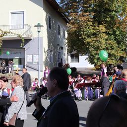 Ein Mann spricht in ein Mikrofon vor einer Menge. Hinter ihm spielt eine Band auf der Straße. Grüne Luftballons sind sichtbar.
