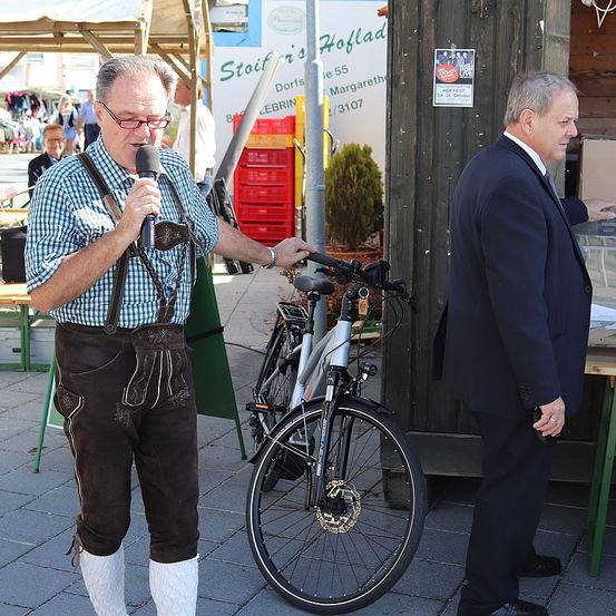 Ein älterer Mann in traditioneller bayerischer Kleidung hält ein Mikrofon in der Hand, während er neben einem Fahrrad steht. Ein Mann in formeller Kleidung steht neben ihm und schaut nach rechts. Dahinter befindet sich eine Holzstruktur mit einem Plakat und einem Tisch.