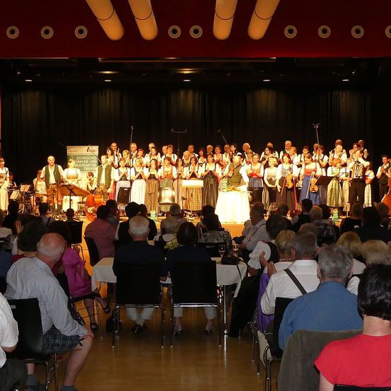 Eine große Gruppe von Musikern tritt auf der Bühne vor einem sitzenden Publikum auf. Die Darsteller umfassen Geiger, Cellisten und Menschen, die andere Streichinstrumente spielen.