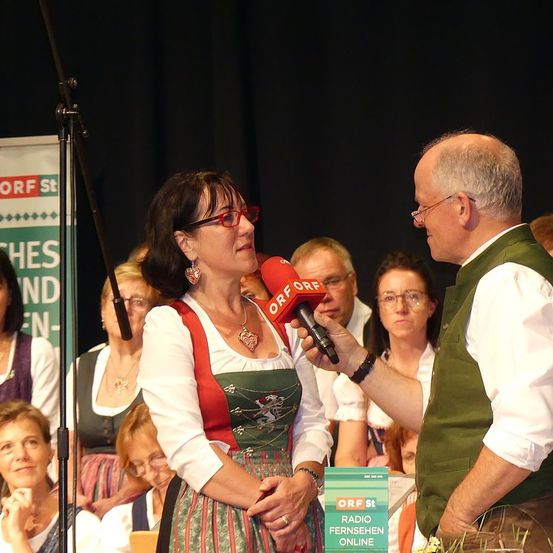 Eine Frau in einem grünen Kleid wird von einem Mann mit einem Mikrofon interviewt, umgeben von Menschen in einem schwach beleuchteten Raum.