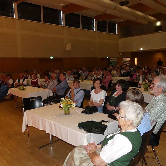 Ein Saal mit Holzboden, gefüllt mit Menschen, die an Tischen sitzen. Viele sind in traditioneller Kleidung gekleidet, und Blumen schmücken die Tische. Die Umgebung deutet auf eine gemeinschaftliche Zusammenkunft oder Veranstaltung hin.
