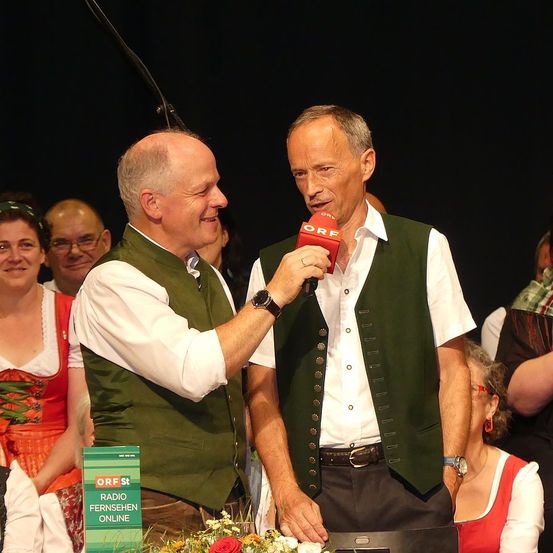 Zwei Männer stehen vor einem Mikrofon, wahrscheinlich bei einer kulturellen Veranstaltung. Der Mann mit dem Mikrofon trägt einen Ring und eine grüne Weste, während der andere Mann in einem weißen Hemd und einer grünen Weste lächelt. Hinter ihnen sind mehrere Menschen anwesend, von denen einige traditionelle Kleidung tragen. Links ist ein Mikrofonstativ zu sehen.