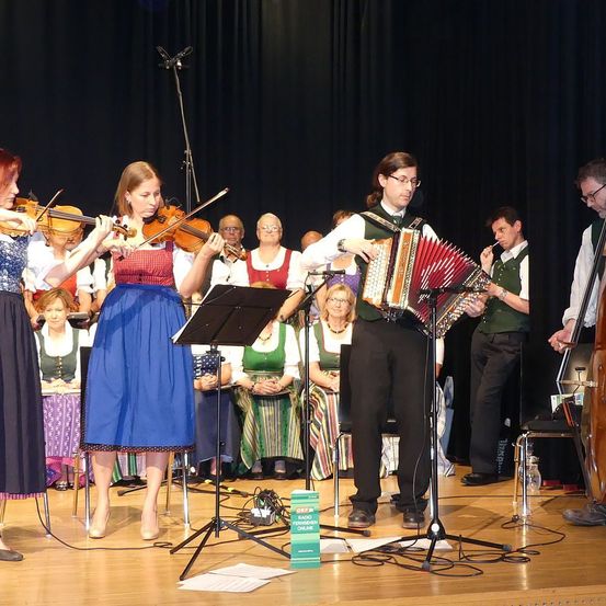 Eine Gruppe von Musikern tritt auf der Bühne auf, darunter zwei Geiger und ein Mann, der ein Akkordeon spielt. Dahinter sitzen mehrere Personen, von denen einige traditionelle Kleidung tragen.