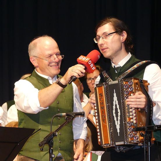 Zwei Männer sind auf der Bühne, einer hält ein Mikrofon und spricht, während der andere ein Akkordeon hält. Sie tragen beide grüne Westen und Brillen. Im Hintergrund sind weitere Personen zu sehen, von denen einige ebenfalls eine Brille tragen.