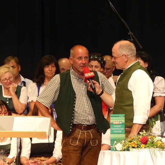 Ein Interview bei einer kulturellen Veranstaltung, zwei Männer mit Mikrofonen, ein Blumenstrauß auf dem Tisch und eine Menge in traditioneller Kleidung.
