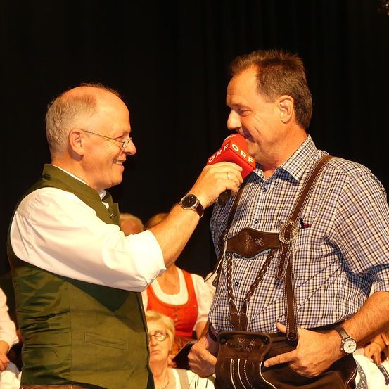 Zwei Männer sind auf der Bühne, einer hält ein Mikrofon und interviewt wahrscheinlich den anderen Mann. Der interviewte Mann trägt eine grüne Weste und eine Brille. Der Interviewer trägt eine Uhr und eine Trägerhose. Ein Publikum ist im Hintergrund zu sehen.