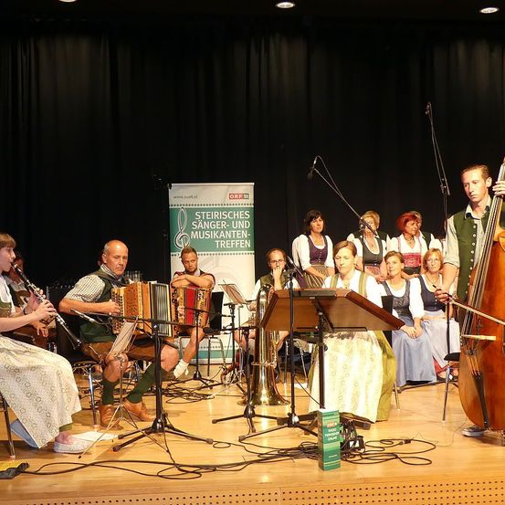 Eine Gruppe von Musikern tritt auf einer Bühne auf und spielt verschiedene Instrumente, mit einem Banner im Hintergrund.