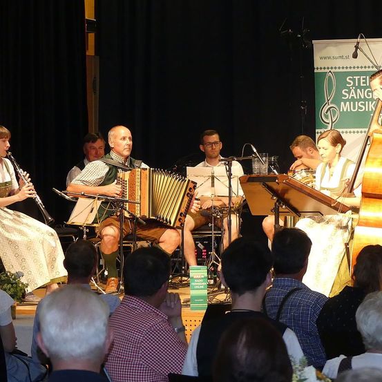 Eine Musikband tritt auf einer Bühne auf, mit einem Banner im Hintergrund. Eine Frau spielt Klarinette, ein Mann Akkordeon und andere spielen Instrumente. Das Publikum beobachtet die Aufführung.