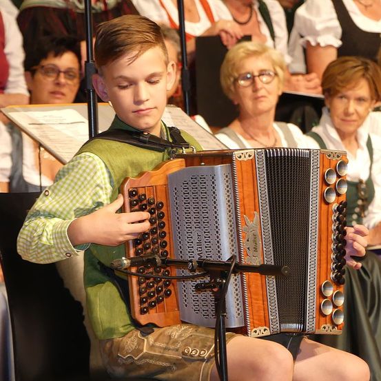 Ein Junge spielt ein Akkordeon, während er vor einem Publikum auftritt. Er trägt ein grünes Hemd und Shorts. Ein Mikrofon ist am Akkordeon befestigt. Dahinter sehen mehrere Personen zu.