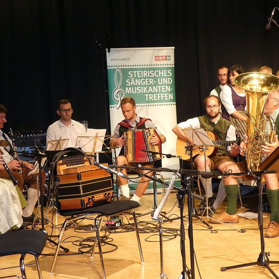 Eine Gruppe von Musikern tritt mit verschiedenen Instrumenten wie Tuba und Akkordeon auf der Bühne auf. Ein Banner hinter ihnen trägt die Aufschrift 'Steierisches Sanger-und Musikanten-Treffen.'