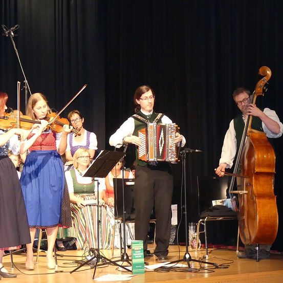 Ein Musikensemble steht auf der Bühne und spielt mit einer Geigerin, einem Akkordeonisten und einem Cellisten. Die Geigerinnen spielen Violinen, während der Akkordeonist und der Cellist mit ihren Instrumenten stehen. Die Bühne ist mit einem schwarzen Hintergrund und einem grünen Buch ausgestattet.