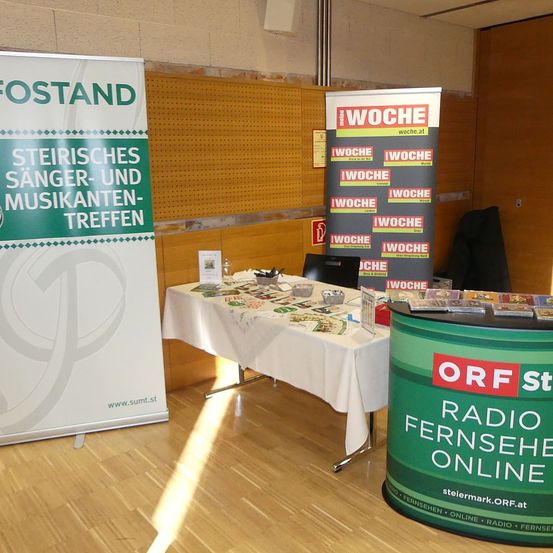 Ein Tisch mit einer weißen Tischdecke, Büchern und einem Computer steht vor zwei Bannern. Die Banner haben die Worte 'Steierisches Sänger- und Musikertreffen' und 'WOCHE'. Der Raum hat einen Holzboden und eine Wand mit einem Aushang.