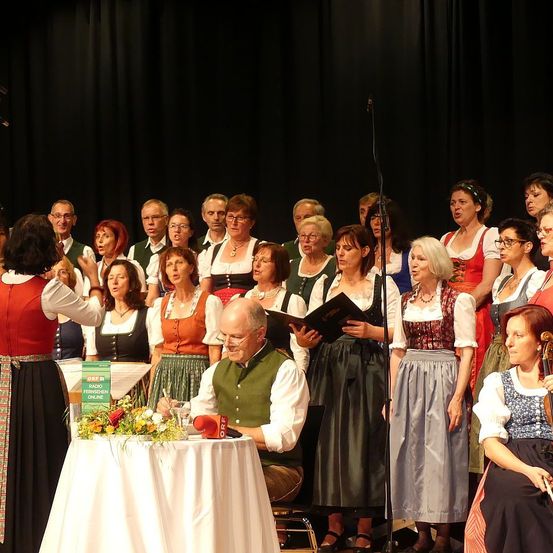 Ein Chor von Menschen in traditioneller bayerischer Kleidung tritt auf der Bühne auf. Ein älterer Mann sitzt vor einem Tisch mit Blumen und hält ein Buch. Eine Frau mit einer Geige steht vor einem Mikrofon.