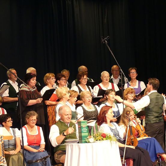 Ein Chor tritt auf der Bühne auf, angeführt von einem Dirigenten. Violinen stehen auf einem Tisch vor einigen Mitgliedern. Sie sind alle in traditioneller Kleidung gekleidet, einige halten Notenblätter in der Hand.