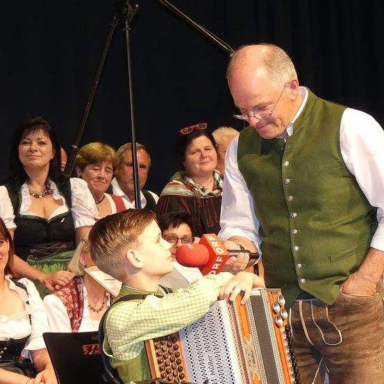 Ein Mann hilft einem jungen Jungen mit einem Akkordeon, während er vor einer Menschenmenge spielt, wahrscheinlich bei einem Festival.