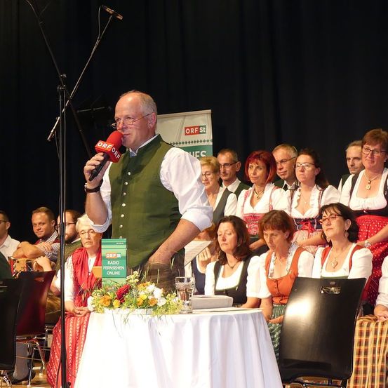 Ein Mann spricht in ein Mikrofon auf der Bühne und trägt eine grüne Weste. Hinter ihm sitzen und stehen mehrere Personen, darunter Frauen in traditionellen Kostümen. Ein schwarzer Stuhl und ein Tisch mit weißer Tischdecke und Blumen sind im Vordergrund zu sehen. Ein Banner mit Text ist im Hintergrund sichtbar.