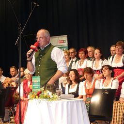 Ein Mann spricht in ein Mikrofon auf der Bühne und trägt eine grüne Weste. Hinter ihm sitzen und stehen mehrere Personen, darunter Frauen in traditionellen Kostümen. Ein schwarzer Stuhl und ein Tisch mit weißer Tischdecke und Blumen sind im Vordergrund zu sehen. Ein Banner mit Text ist im Hintergrund sichtbar.