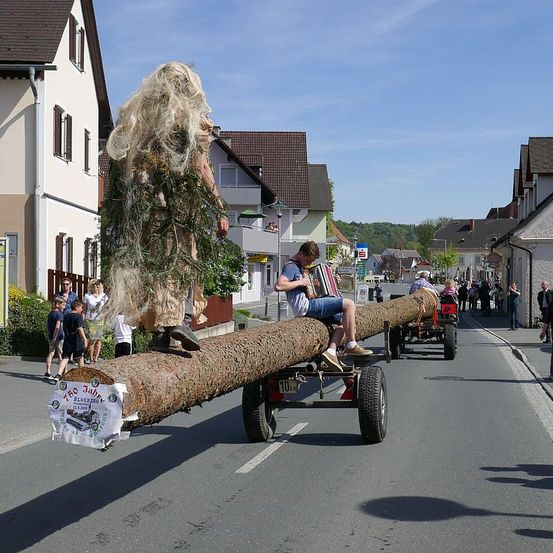 Ein Mann spielt Akkordeon, während er auf einem langen Baumstamm sitzt, der von einem Anhänger gezogen wird. Im Hintergrund sind Menschen und Gebäude zu sehen.
