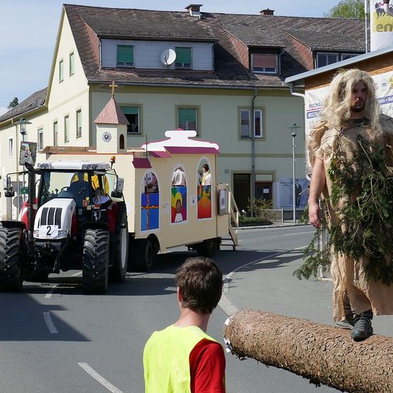 Ein Traktor, der mit farbenfrohen Gemälden verziert ist, fährt auf der Straße. Eine Person sitzt im Inneren des Traktors. Ein Junge steht davor und sieht ihn an. Ein Mann steht an der Seite, gekleidet in einem Kostüm.
