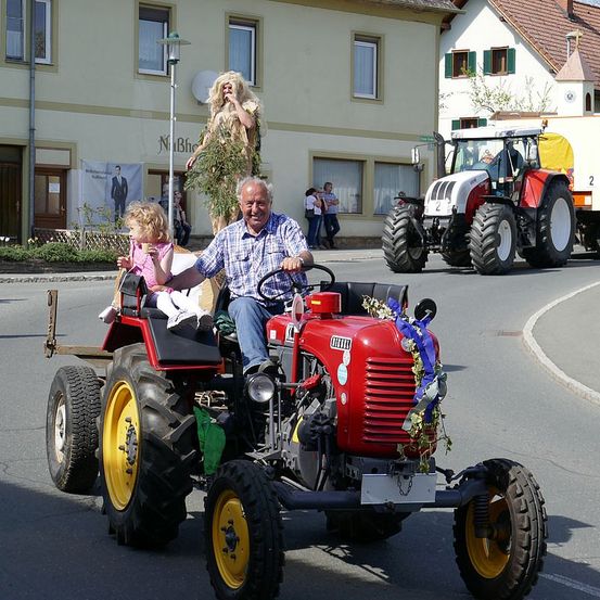 Ein älterer Mann fährt einen roten Traktor mit einem kleinen Mädchen, während ein anderer Traktor dahinter fährt. Eine große Baumfigur steht in der Nähe.