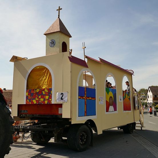 Ein kirchenförmiger Umzugswagen mit Uhrenturm, gekennzeichnet mit '2', steht auf der Straße. Er hat farbenfrohe Fenster und Menschen sitzen darin. Ein Traktor zieht ihn.