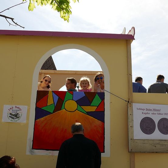 Eine Gruppe von Menschen schaut aus einem gelben Stand heraus, mit einem farbenfrohen abstrakten Gemälde dahinter. Ein Banner mit zwei Münzen ist an der Wand angebracht.