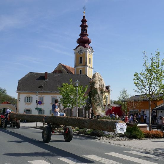 Ein großer Baumstamm wird an einem sonnigen Tag von einem Traktor gezogen. Eine Person steht auf dem Stamm, während ein Mann daneben sitzt und ein Musikinstrument spielt. Im Hintergrund ist ein Kirchturm zu sehen.