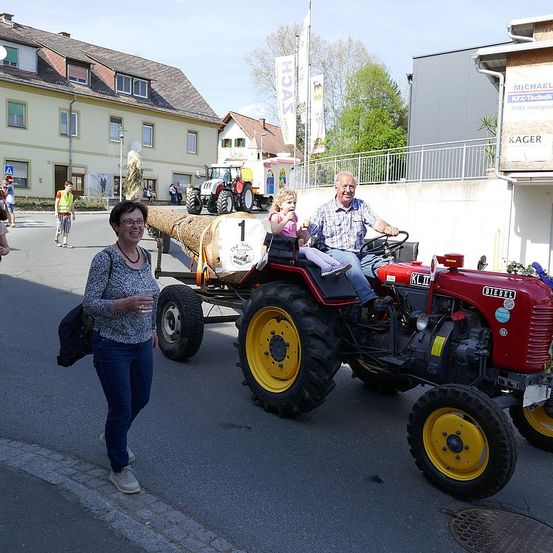 Ein älterer Mann fährt einen roten Traktor, während ein kleines Mädchen auf seinem Schoß sitzt. Eine Frau geht neben ihnen. Hinter ihnen befindet sich ein großer Baumstamm und ein Gebäude mit einem Schild, das 'Kager' sagt.