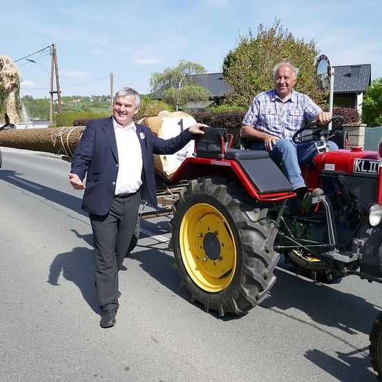 Zwei Männer sind auf einem Traktor bei einer Parade. Ein Mann fährt und der andere steht neben dem Traktor. Ein großer Baumstamm ist auf der Rückseite des Traktors. Dahinter befindet sich ein Gebäude mit einer Steinmauer.
