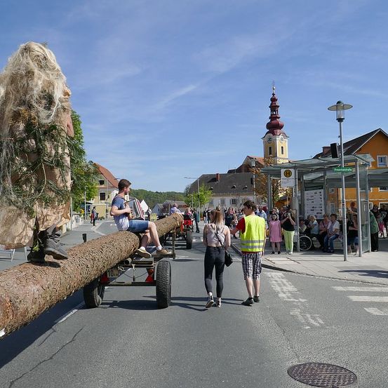 Ein Umzug an einem sonnigen Tag mit einem Mann, der Akkordeon auf einem Baumstamm spielt, während andere am Straßenrand gehen und sitzen. Eine hohe Turmspitze ist in der Ferne sichtbar.