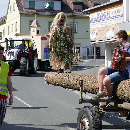 Ein Mann, der als Baum gekleidet ist, steht auf einem Baumstamm, während ein anderer Akkordeon spielt. Dahinter steht ein Traktor und ein Gebäude mit einem Schild mit der Aufschrift ZACH.