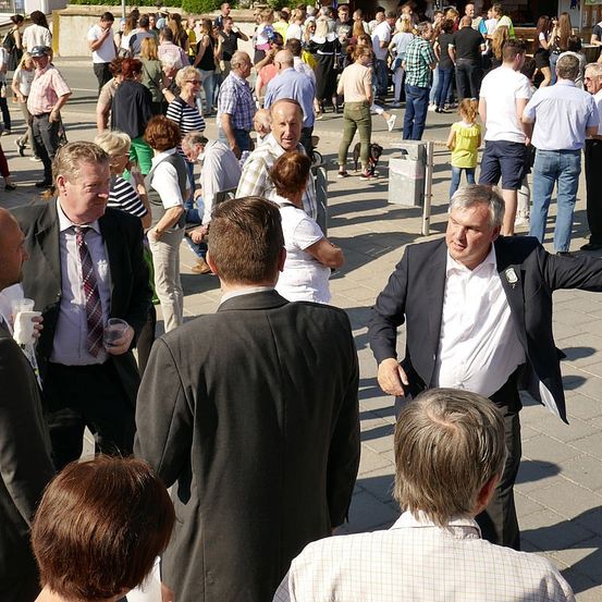 Eine Menschenmenge, größtenteils mittleren Alters, versammelt sich auf einem Außengelände. Einige tragen formelle Kleidung, während andere in Freizeitkleidung sind. Ein Mann in einem Anzug gestikuliert mit seiner Hand.