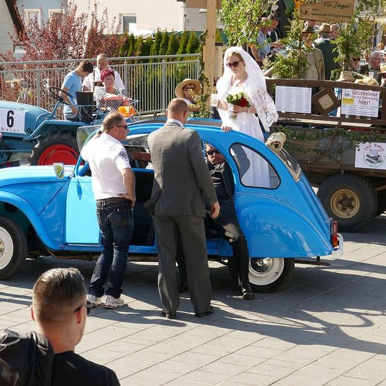 Eine Braut in einem weißen Kleid hält einen Blumenstrauß und steht auf einem blauen Auto. Mehrere Leute stehen um sie herum, darunter zwei Männer in Anzügen. Hinter dem Auto steht ein Traktor mit der Nummer 6.
