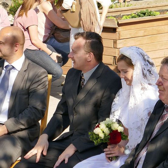 Eine Gruppe von Menschen sitzt draußen zusammen. Ein Mann und eine Frau halten Händchen und schauen sich an. Die Frau trägt ein weißes Kleid und eine Schleier.