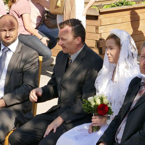 Eine Hochzeitszeremonie mit einer Braut und einem Bräutigam, die mit ihren Familien sitzen. Die Braut hält einen Strauß weißer und roter Blumen. Hinter ihnen sitzen und stehen andere, einschließlich einer Frau mit einer Handtasche.