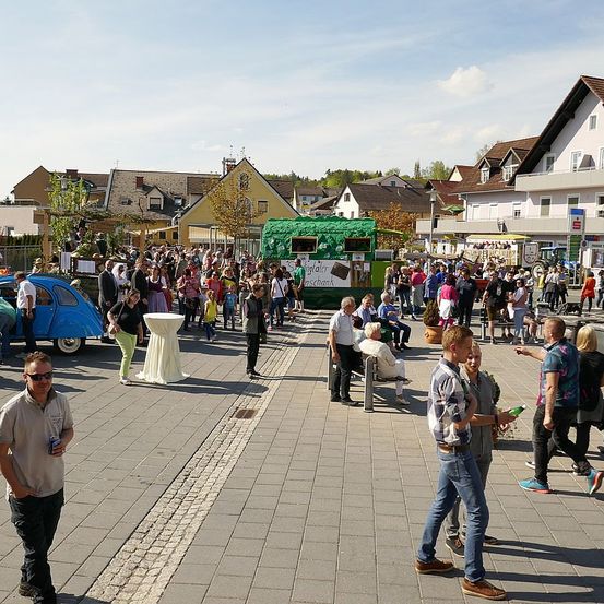 Ein geschäftiger Stadtplatz mit vielen Menschen, einem grünen Anhänger, Autos und Häusern. Einige Leute gehen, während andere auf Bänken sitzen.