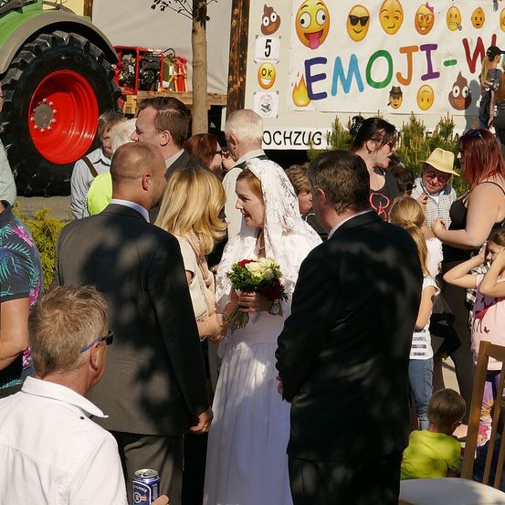Eine Hochzeitszeremonie findet im Freien statt, an der eine große Gruppe von Menschen teilnimmt. Die Braut trägt ein weißes Kleid, hält Blumen und der Bräutigam steht neben ihr. Ein Traktor ist im Hintergrund geparkt.