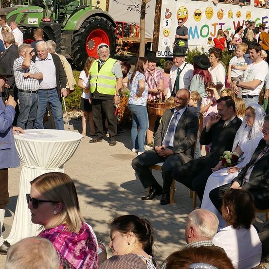 Eine große Menschenmenge versammelt sich im Freien, einige sitzen auf Stühlen, andere stehen, in der Nähe eines Traktors und einer Banner mit Emojis und Text.