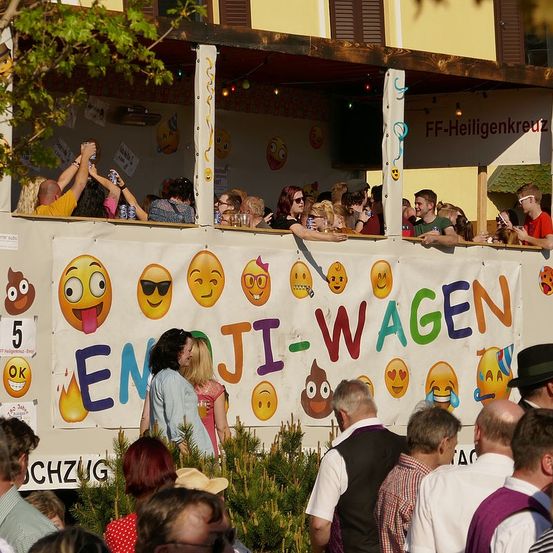 Eine Gruppe von Menschen beobachtet eine Veranstaltung im Freien, möglicherweise eine Parade, von einer erhöhten Position aus. Sie stehen vor einem dekorierten Wagen mit dem Wort Emoji. Der Wagen hat verschiedene Emojis und Text darauf geschrieben. Sie tragen alle Sonnenbrillen und einige von ihnen halten Dosen. Vor dem Wagen befinden sich Bäume und Pflanzen.