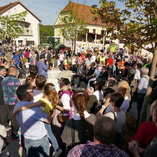 Eine große Menschenmenge versammelt sich im Freien vor einem Gebäude, einige sitzen, andere stehen, mit Traktoren und Bäumen im Blick. Die Menschen tragen Freizeit- und traditionelle Kleidung.