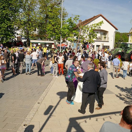 Eine große Menschenmenge versammelt sich auf einer Straße in einer Stadt. Viele tragen Freizeitkleidung, und einige haben Taschen dabei. Es gibt Essensstände und einen Traktor in der Nähe. Bäume und Häuser säumen die Straße.