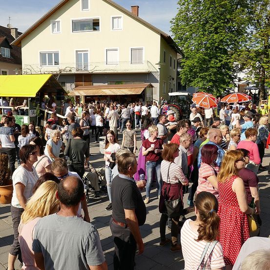 Eine große Menschenmenge versammelt sich im Freien, vermutlich auf einem Markt. Einige Personen stehen, während andere unter Regenschirmen sitzen. Im Hintergrund befinden sich Stände und Gebäude.