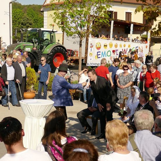 Eine Menschenmenge bei einer Veranstaltung im Freien. Ein Mann in einem blauen Anzug spricht in ein Mikrofon. Ein anderer Mann in einem schwarzen Anzug sitzt auf einem Stuhl. Im Hintergrund steht ein Traktor und ein Baum.